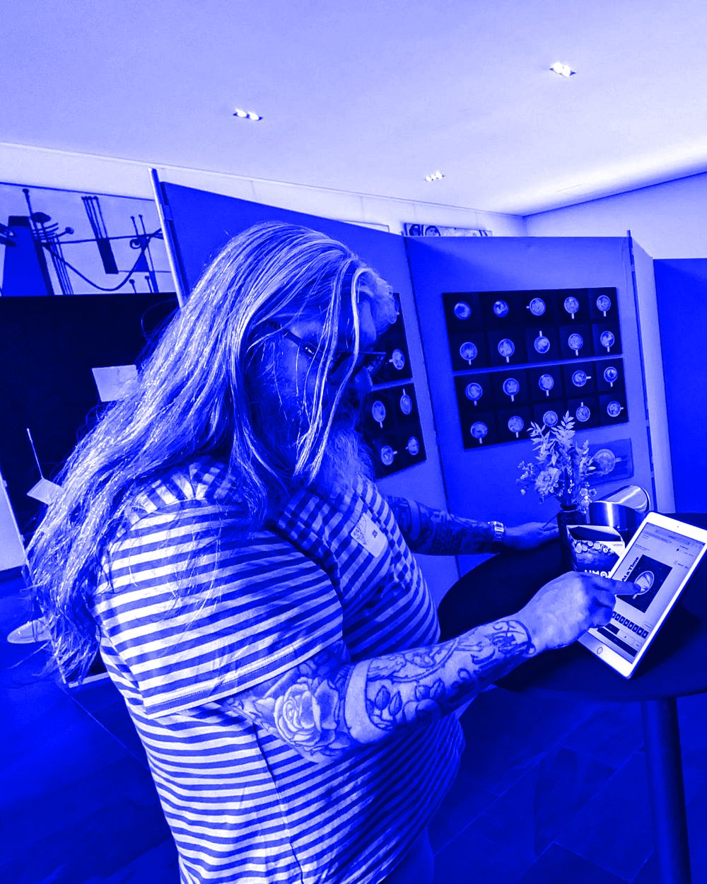 A visitor in a striped shirt painting on the iPad at a cocktail table, the panels of coffee paintings behind him
