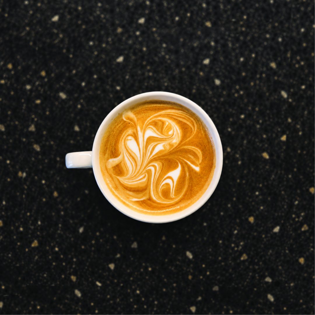 Coffee painting Nº 08: a white cup seen from above on a dark speckled counter, the foam pulled with a teaspoon into dense, intricate marbling